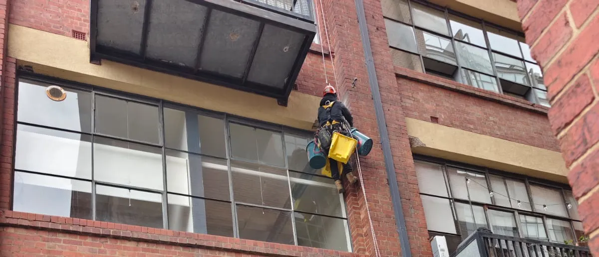 Rope-access technician cleaning windows on a brick building facade with suspended gear and buckets.